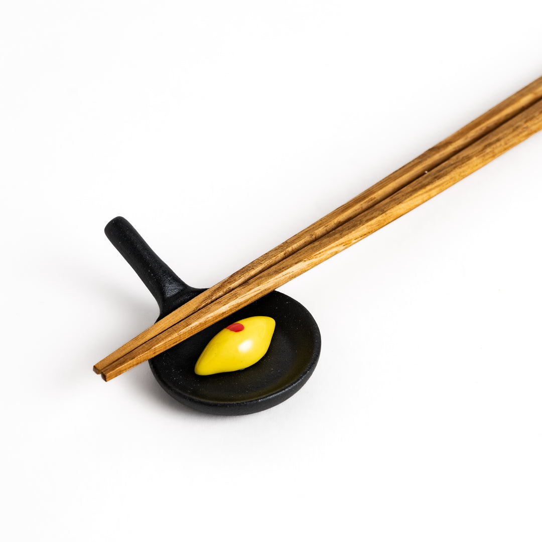 Wooden chopsticks resting on a unique chopstick rest shaped like a miniature frying pan with a Japanese omelette rice.