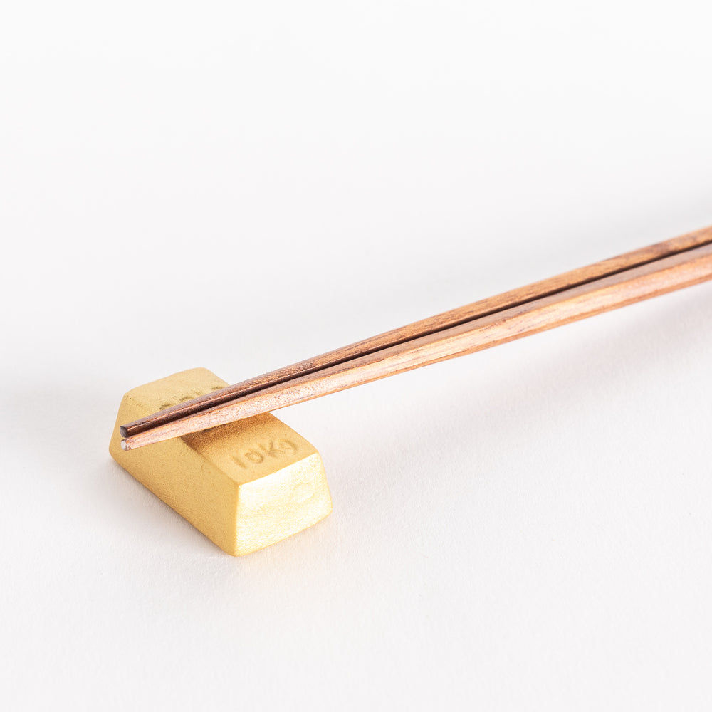 A close-up view of a gold bar-shaped ceramic chopstick rest holding chopsticks on its flat surface.