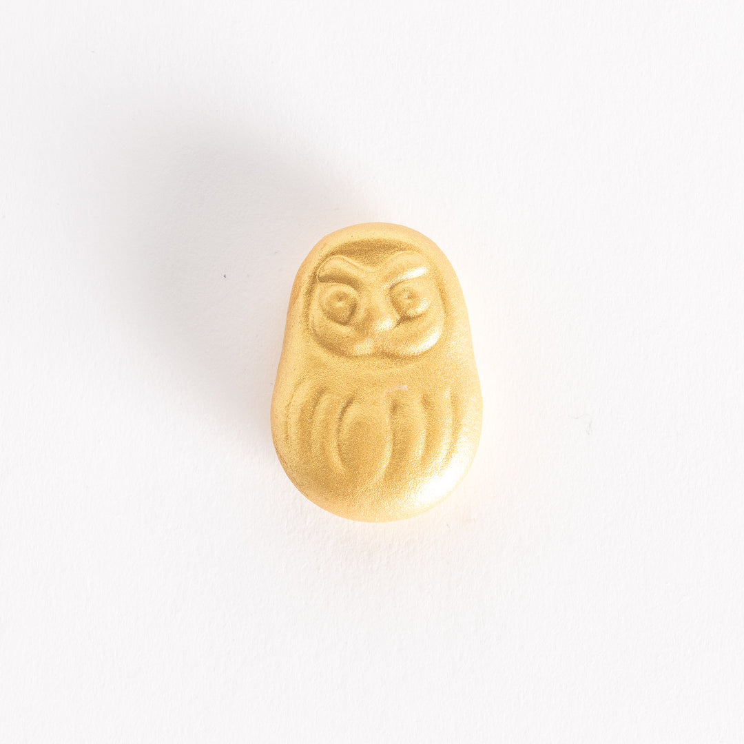 A gold ceramic chopstick rest shaped like a Daruma doll, featuring a smooth, detailed design.