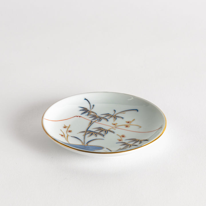 A delicate round sauce dish featuring bamboo, a flowing river, and a distant mountain in shades of blue and orange, finished with a gold rim.