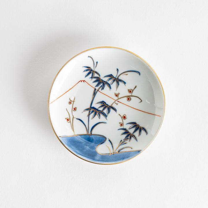 A round sauce dish with a delicate scene of bamboo, a flowing river, and a distant mountain in blue and orange tones. The dish is finished with a gold rim.