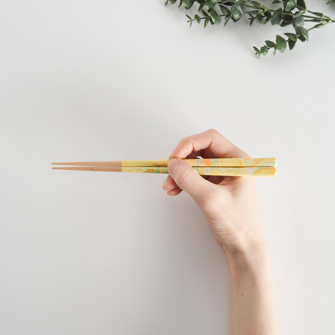 A hand holding the marbled chopsticks with hues of yellow, orange, and green, showcasing their elegant design and functionality.