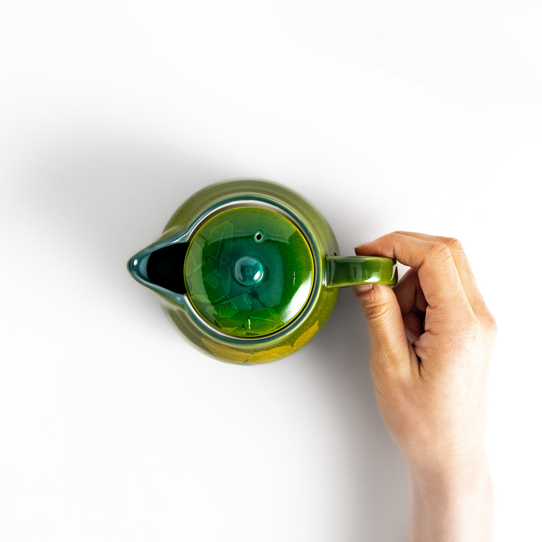 Hand gracefully holding the handle of a green ceramic teapot with a matching lid, viewed from above. 