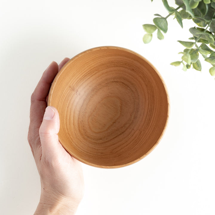 Hand holding the cherry wood bowl, highlighting its smooth interior and natural grain pattern. Green leaves are visible in the background.