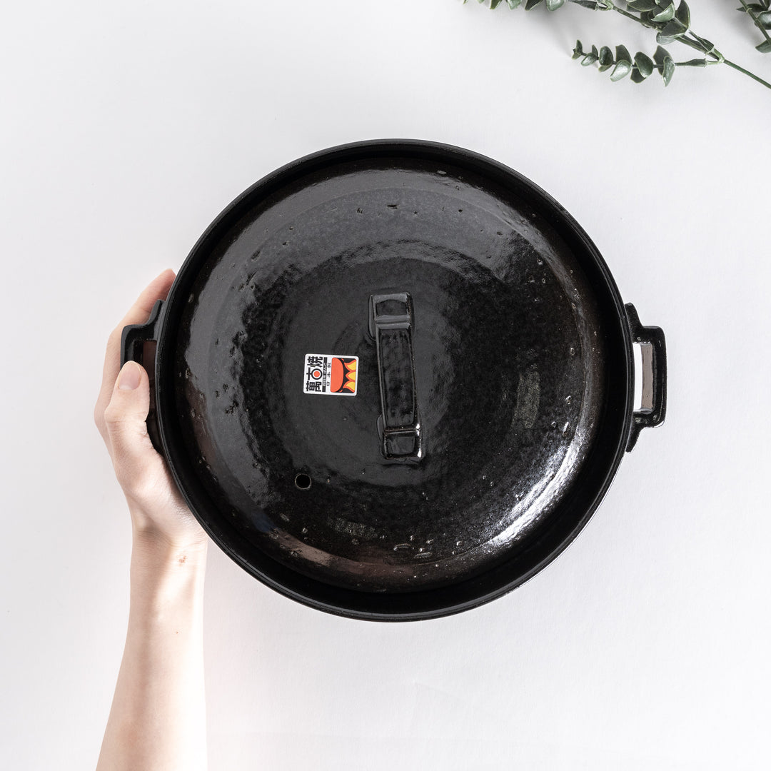 Top-down view of a glossy black donabe pot held in hand, highlighting the lid's square handle.