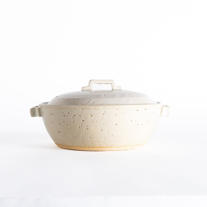 Side view of an off-white donabe pot with a rounded design, soft speckled details, and sturdy handles.