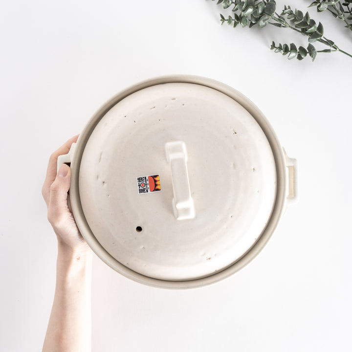 Top-down view of an off-white donabe pot held in hand, emphasizing the square handle and smooth lid.