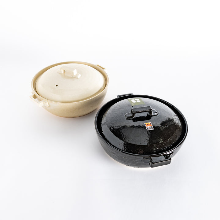 Two donabe pots side by side: one with a glossy black glaze and the other in a smooth, off-white finish, both featuring square handles and lids.