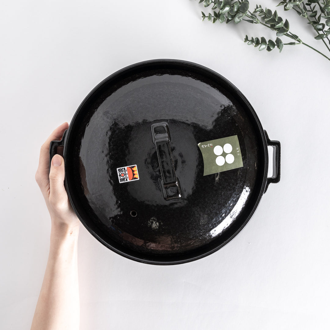 Top-down view of a glossy black donabe pot held in hand, highlighting the lid's square handle.