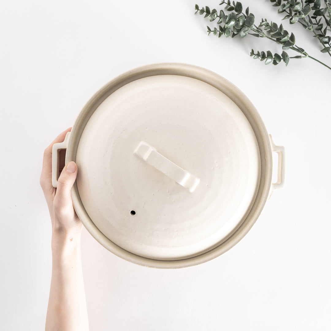 Top-down view of an off-white donabe pot held in hand, emphasizing the square handle and smooth lid.