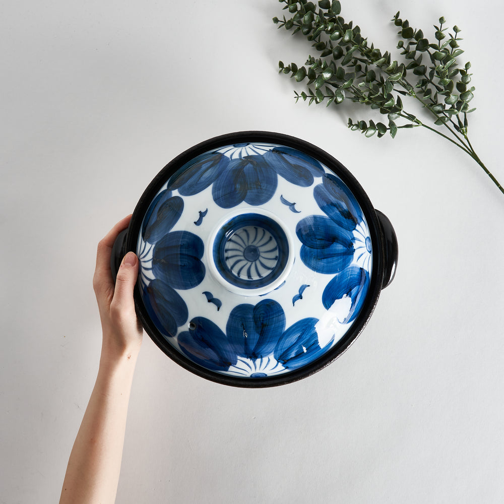 A hand gently holding the donabe, highlighting its indigo floral design and intricate details on the lid.