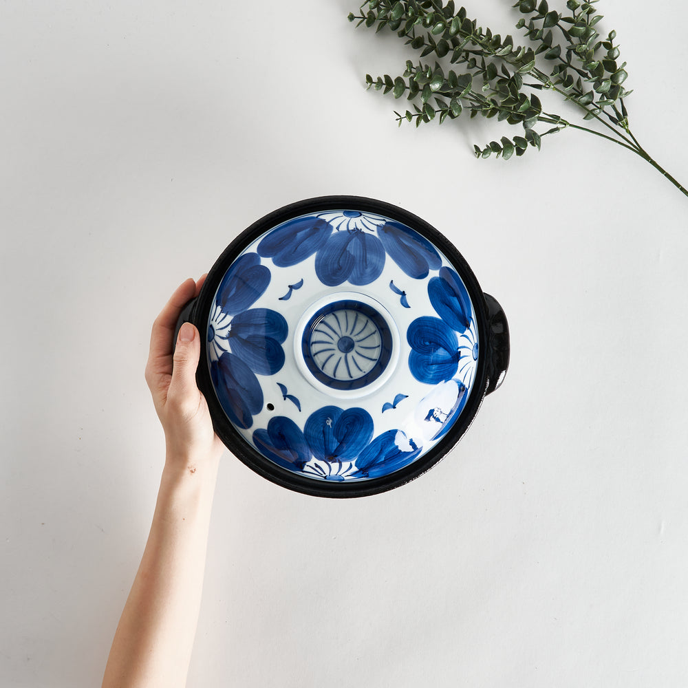 A hand holding the donabe from above, showcasing the detailed blue flower design on the lid with a central vent hole.