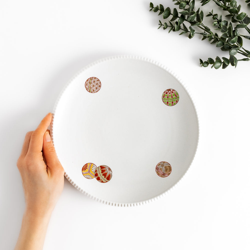 Hand holding a white dinner plate featuring colorful temari ball designs.