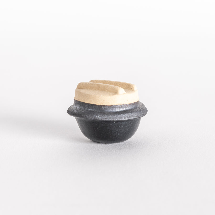 Miniature kamameshi pot-shaped chopsticks rest with a beige lid and black base.