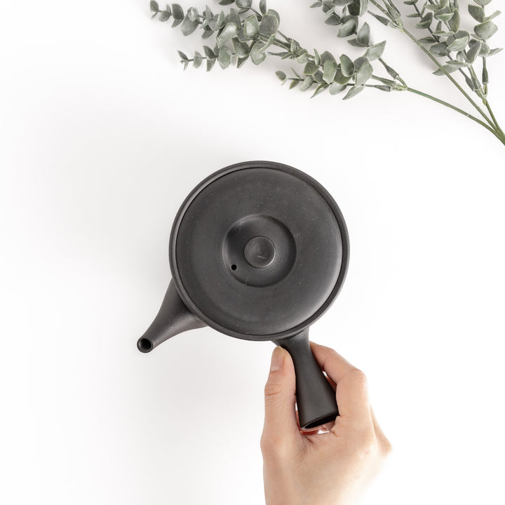 Top-down view of the black teapot with a hand on the handle, showcasing its functional and ergonomic design.