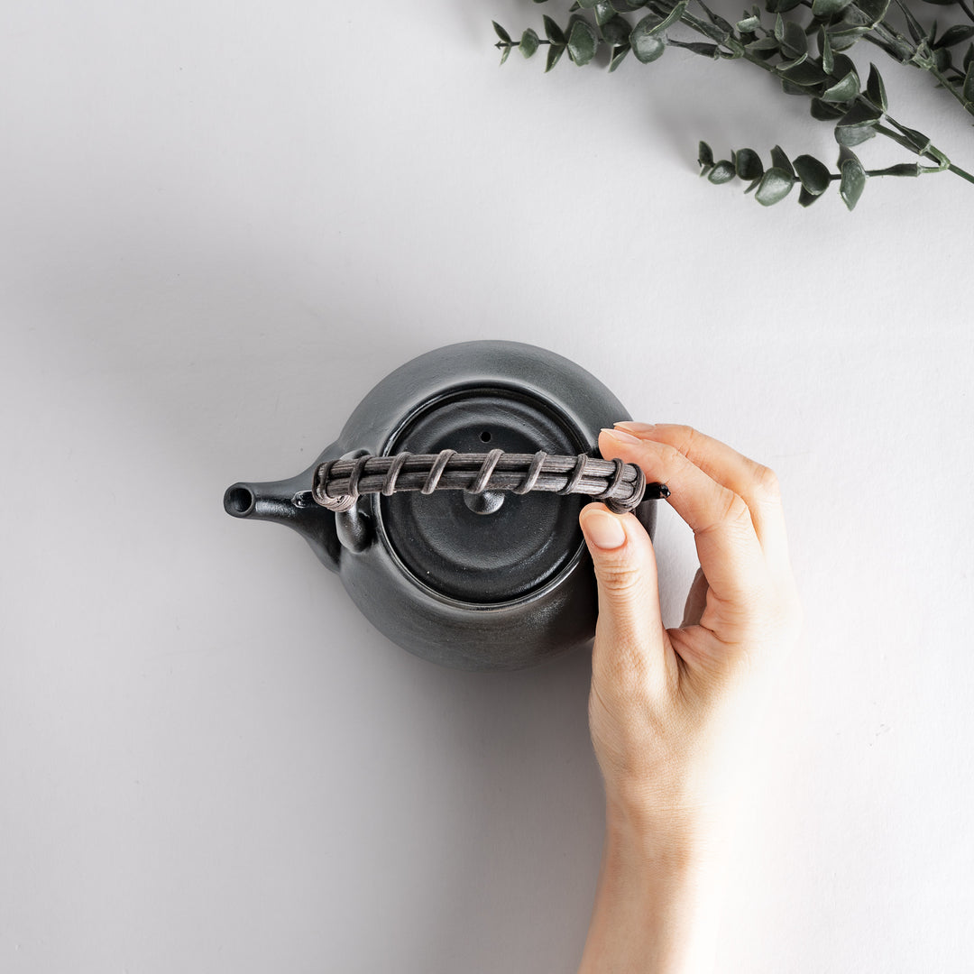 Top view of the matte black teapot held by a hand, highlighting the braided handle and smooth surface of the teapot.