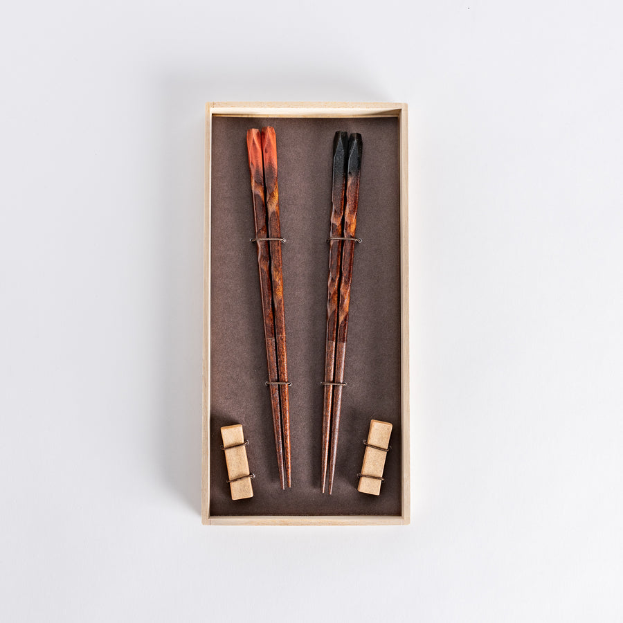 Boxed set of dark brown chopsticks with textured designs and matching rests, placed neatly in a wooden box on a dark background.