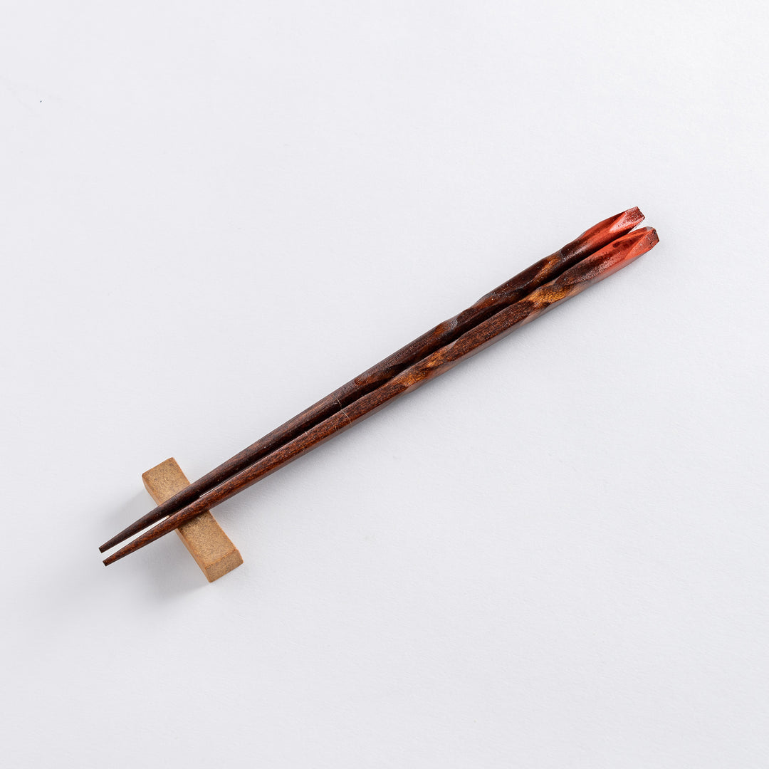 A dark wooden chopsticks with finely tapered tips resting on a simple rectangular chopstick rest, showcasing a smooth, elegant finish.