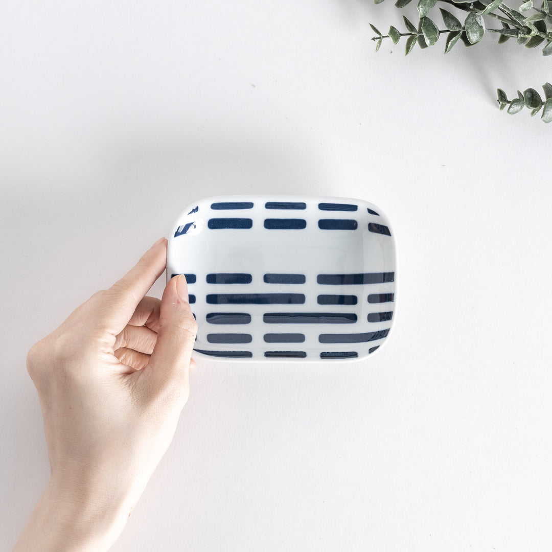 A hand holding a rectangular sauce dish from above, with blue striped patterns on the surface.