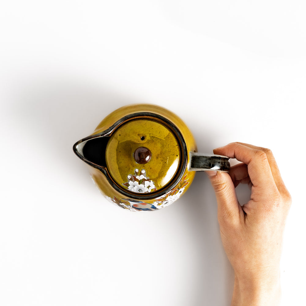 Hand holding the handle of a yellow ceramic teapot with a bird and flower design, viewed from above. The teapot features a matching lid and a comfortable grip.