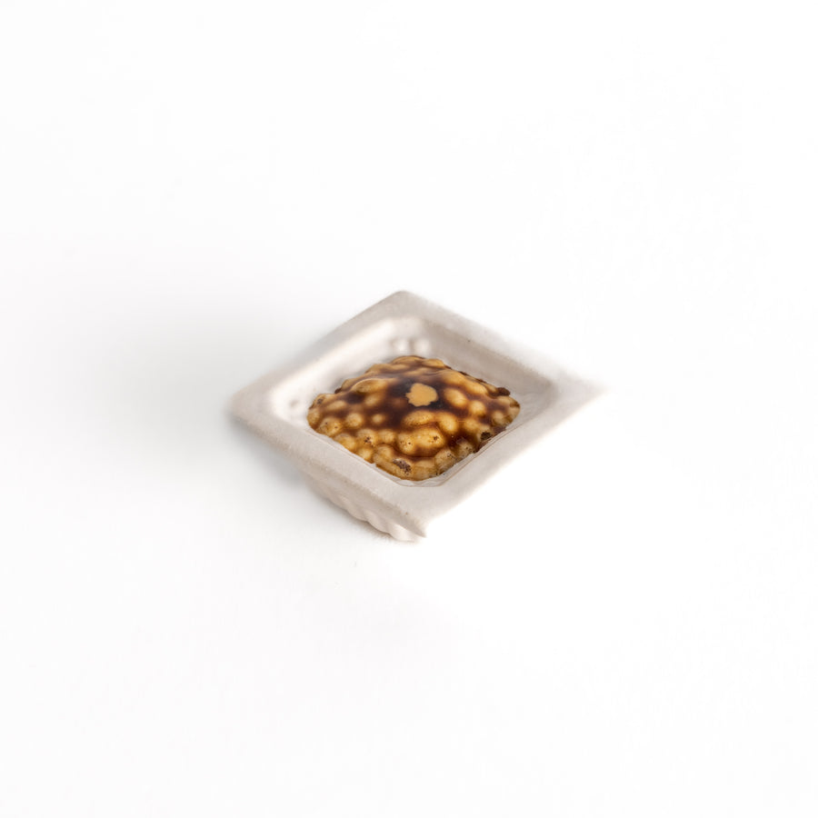 A ceramic chopstick rest in the shape of a square bowl, featuring a textured natto bean design in brown and white tones.