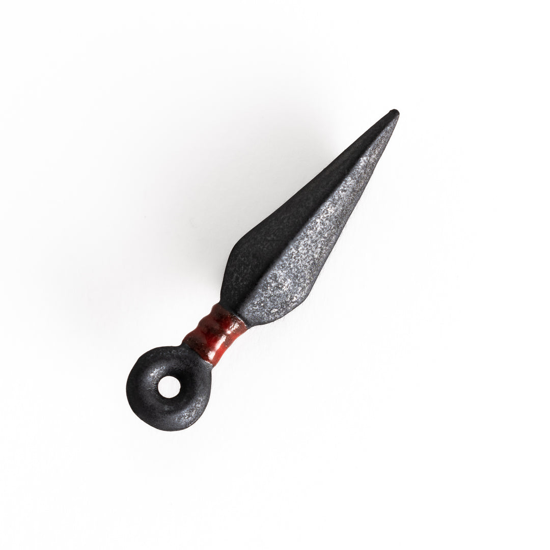 A kunai-shaped ceramic chopstick rest in black with a red accent, featuring a textured matte finish and loop detail at the handle's end.