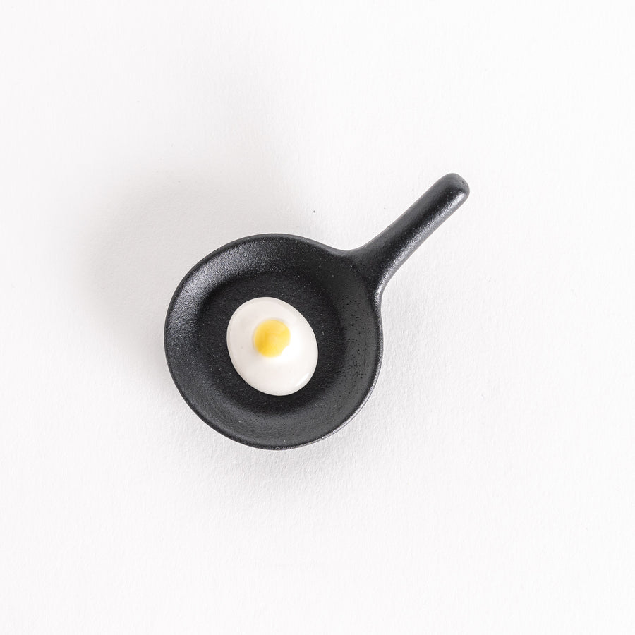 Black frying pan-shaped chopsticks rest with a white fried egg design featuring a yellow yolk in the center.