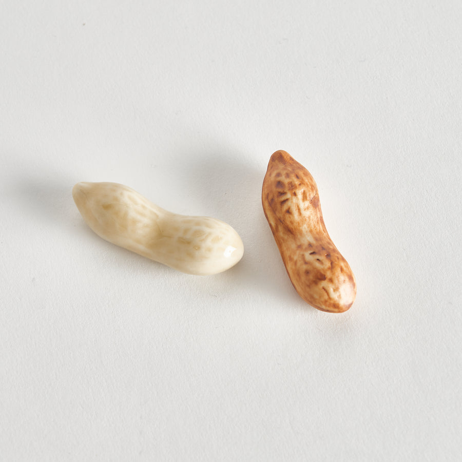 A pair of chopstick rests shaped like peanuts, one with a pale cream color and the other with a brown, roasted appearance, showcasing detailed texture.