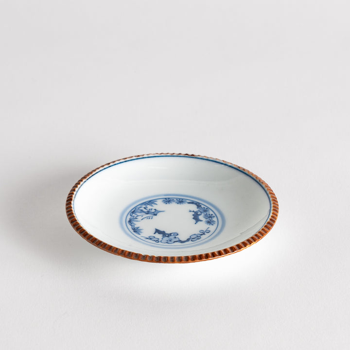 A small ceramic plate with a central blue floral motif surrounded by a white background, framed by a copper-colored textured rim.