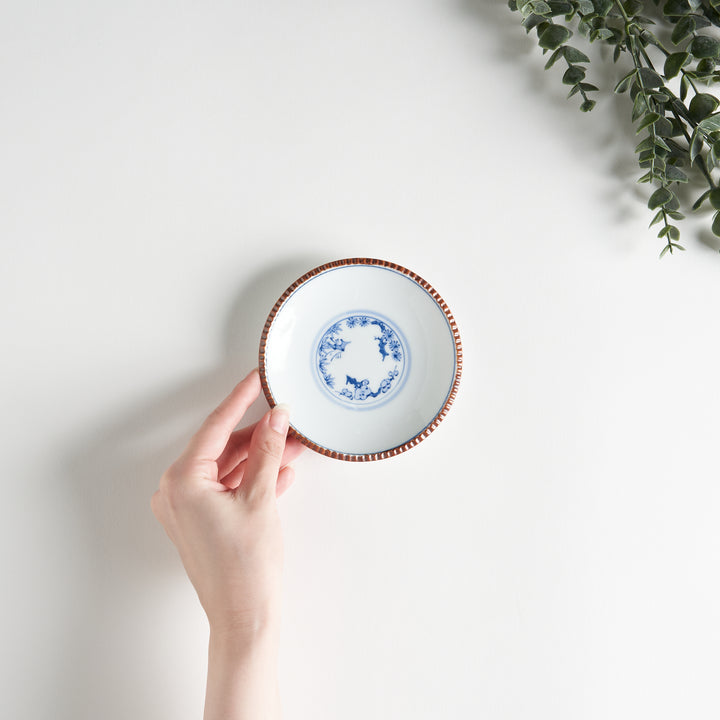 A small ceramic plate with a central blue floral motif surrounded by a white background, framed by a copper-colored textured rim.