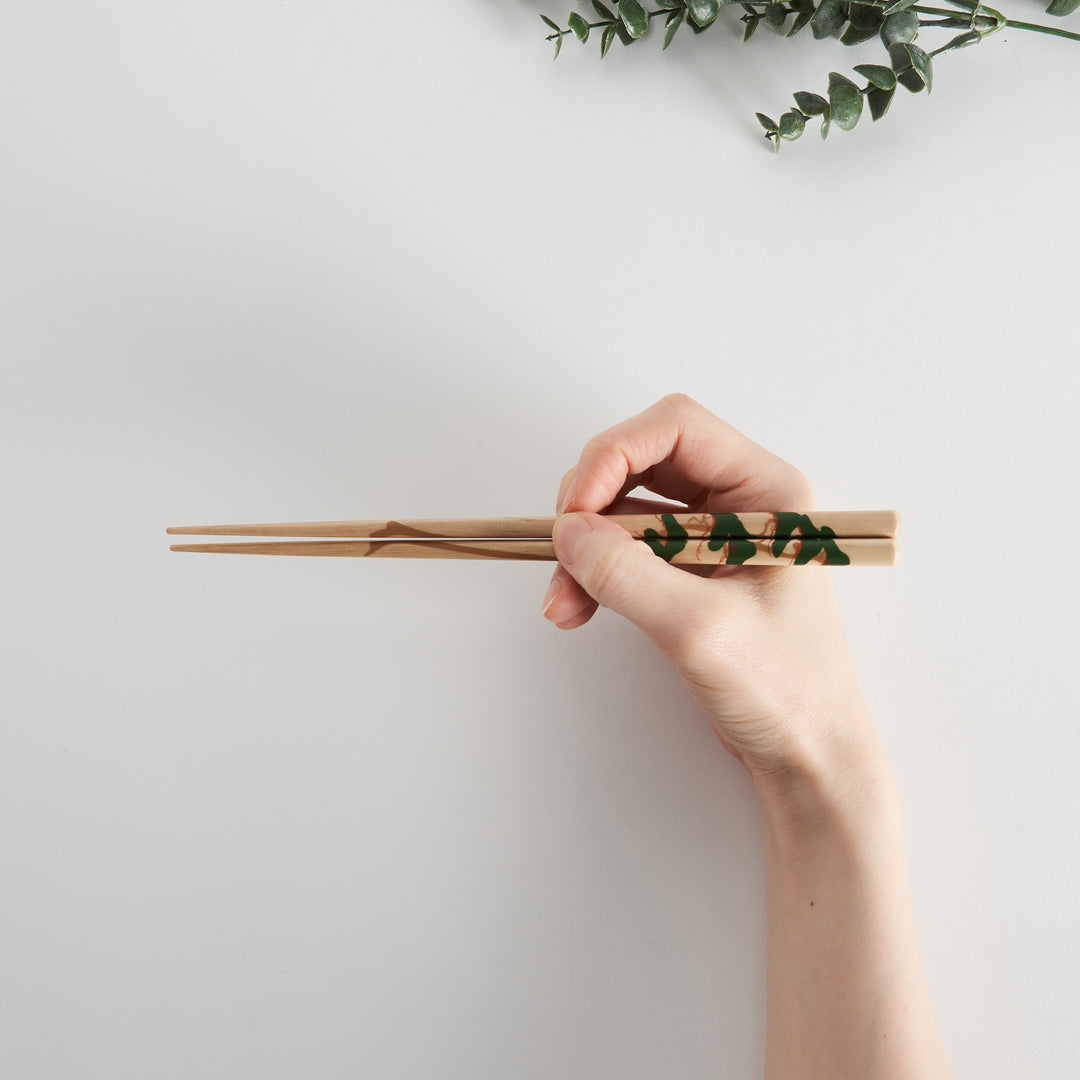 A hand gripping a pair of chopsticks with an intricate green and brown leaf design, emphasizing their elegant and functional design.