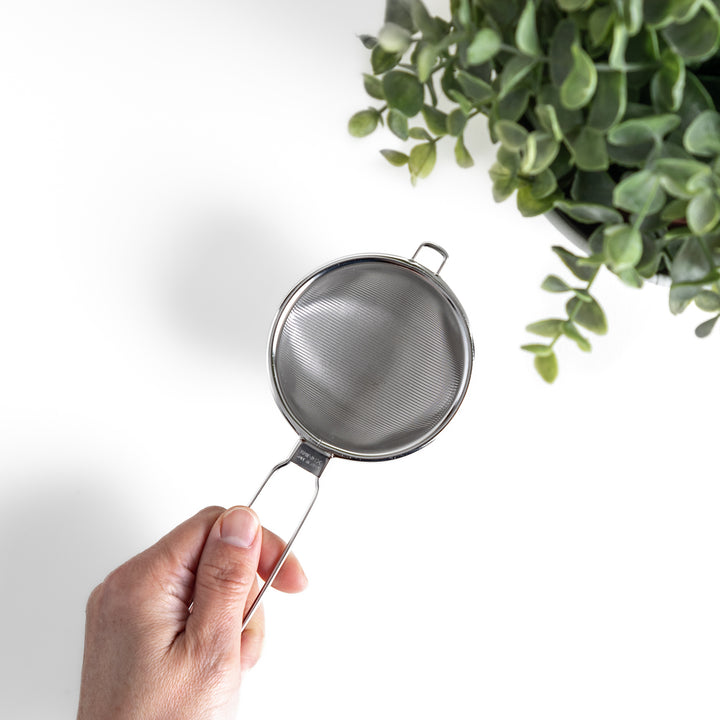 Hand holding a stainless steel tea strainer with a fine mesh and convenient handle, a versatile kitchenware tool for brewing loose leaf tea.