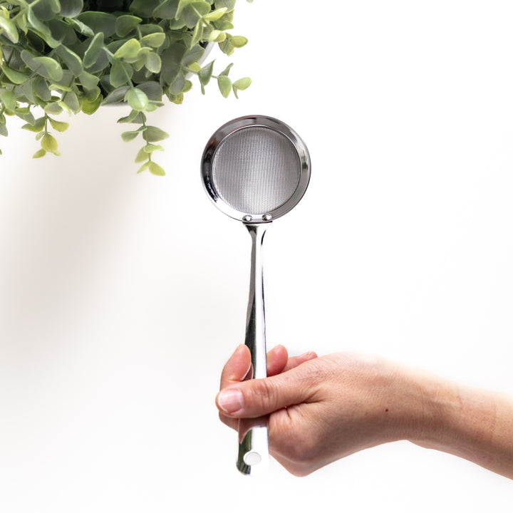 Hand holding a stainless steel skimmer with a fine mesh net and a long handle, a useful kitchenware tool for removing impurities from liquids.