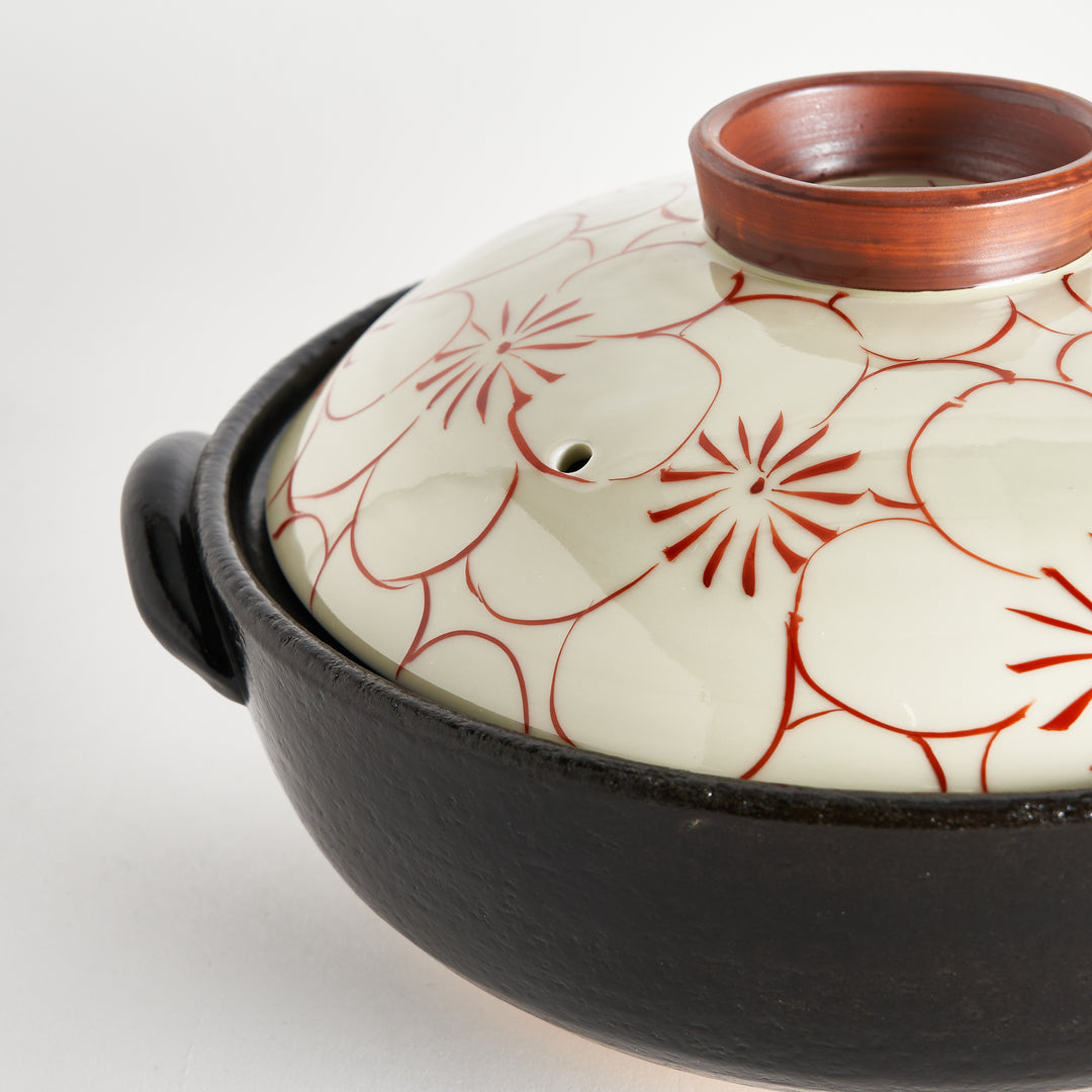 A close-up of the donabe's black base and the lid decorated with a delicate red flower design and a small ceramic lid top.