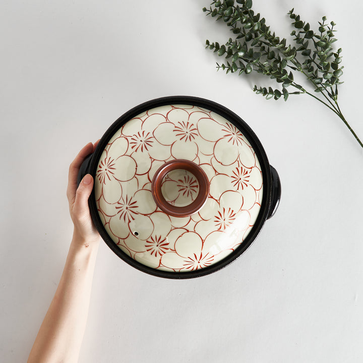 A hand holding the donabe from above, showcasing its red floral lid design and black base, with a sprig of greenery beside it.