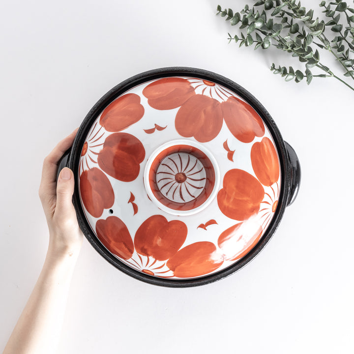 Hands holding the lid of a donabe pot with bold red floral patterns, set on a portable gas stove, with small side dishes around.