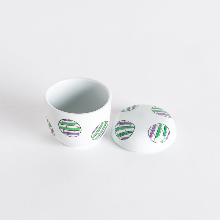 Open white bowl revealing a lid adorned with alternating green and purple striped circles.