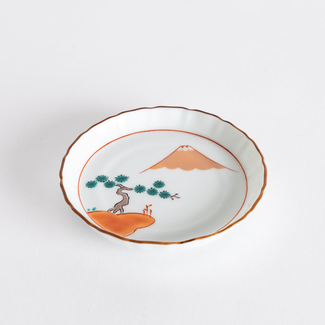 Top view of a round dish adorned with a scenic landscape of Mount Fuji and a red pine grove, featuring a delicate scalloped edge.