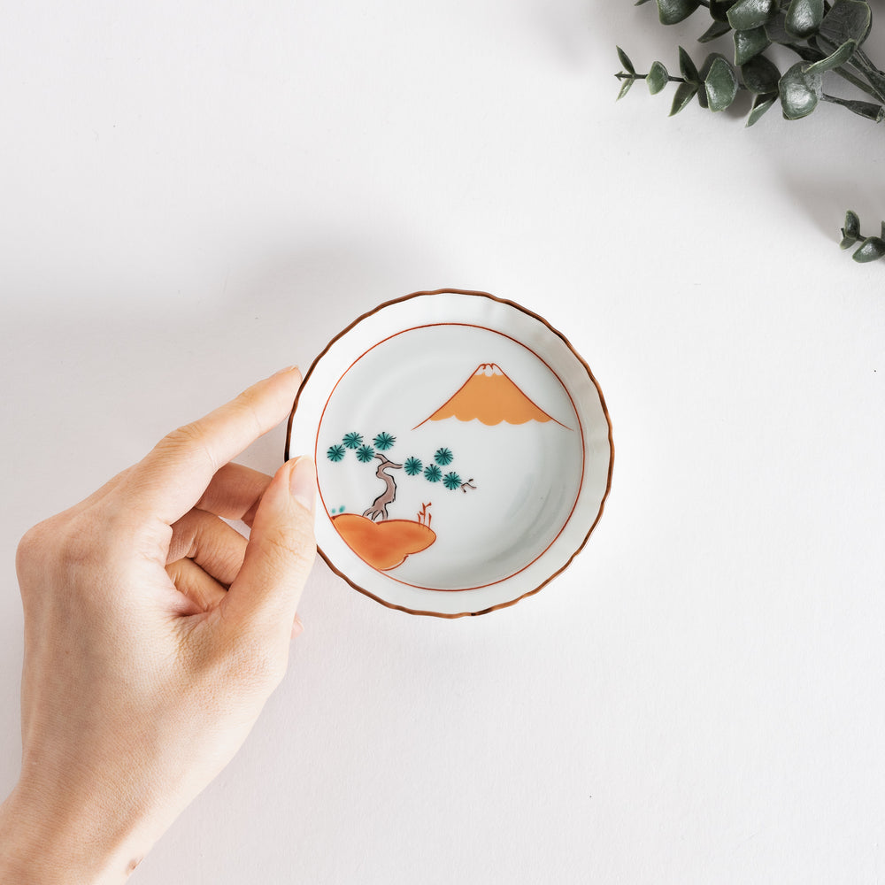 Hand holding a decorative dish with Mount Fuji and a red pine grove design, set against a minimalistic background.