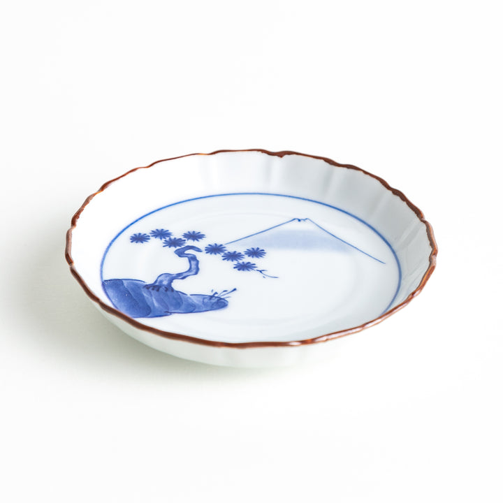 Angled view of a porcelain sauce dish displaying its slightly raised rim and blue Mount Fuji and pine tree motif, with a brown accent on the edge.