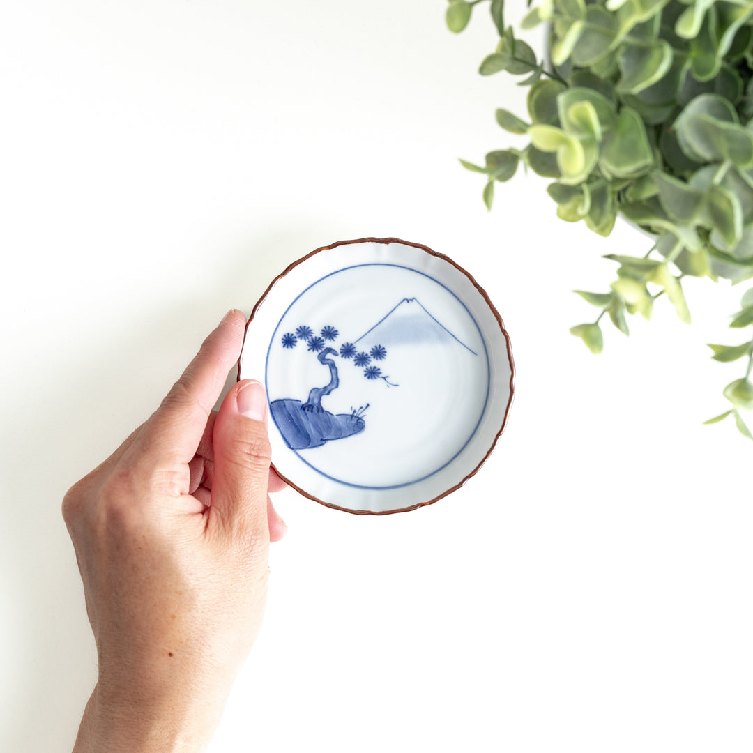 Hand holding a porcelain sauce dish, showcasing its size and intricate blue Mount Fuji and pine tree design with brown edge detailing.