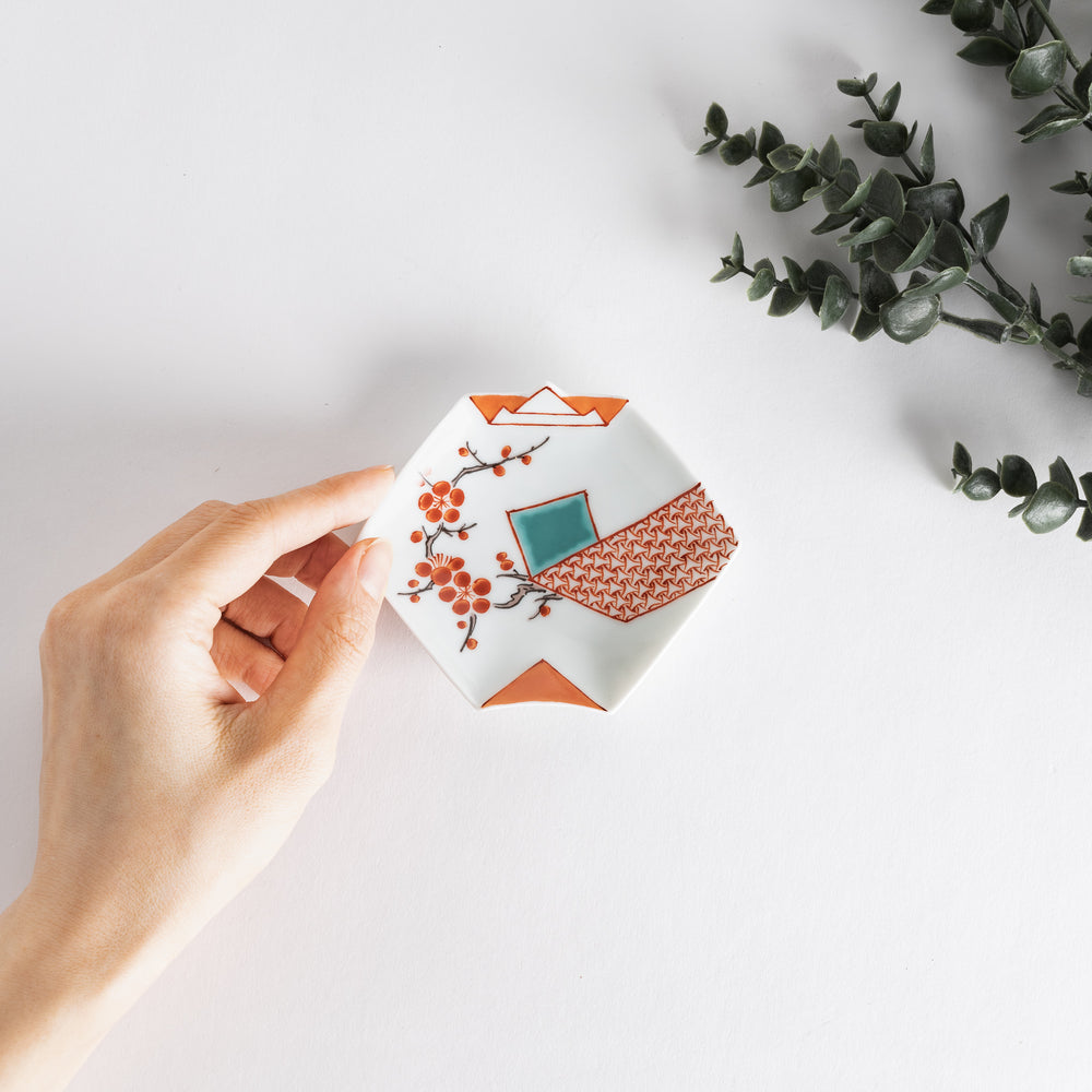 A hand gently holding a square sauce dish, decorated with orange plum blossoms and geometric patterns in orange and blue.