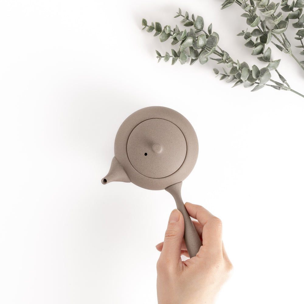 Top-down view of the gray teapot with a hand gently holding the side handle, emphasizing its ergonomic design.