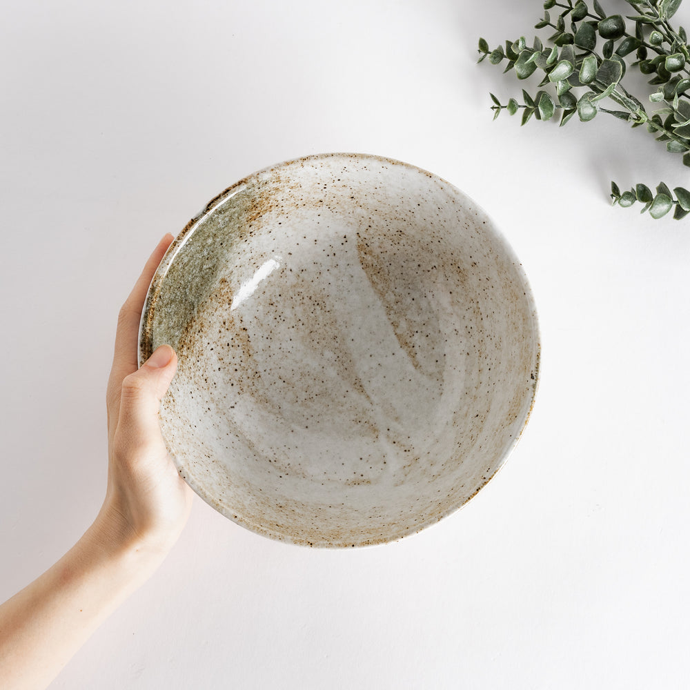 Hand holding a rustic earth-tone ramen bowl with a speckled glaze and subtle green and brown accents.