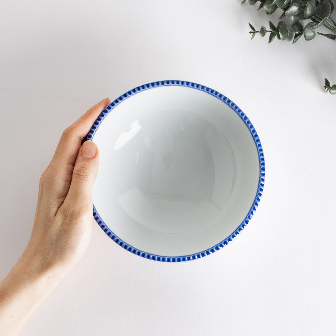 A hand holding the cereal bowl, showcasing its smooth white interior and blue scalloped edge.