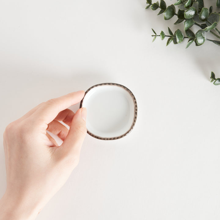 A hand holding a small oval sauce dish, showcasing its scalloped edge and brown dotted rim.