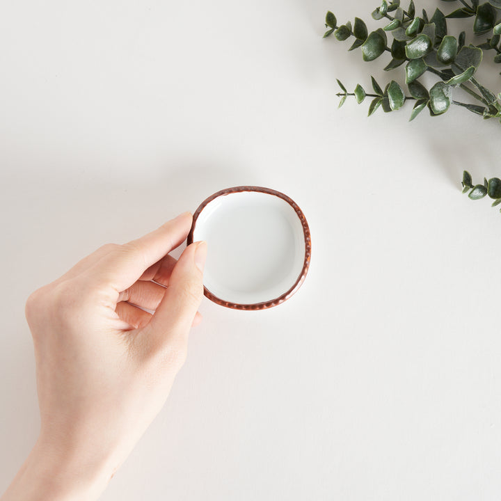 Hand holding a small oval sauce dish, showcasing its smooth white interior and the scalloped olive-colored edge.