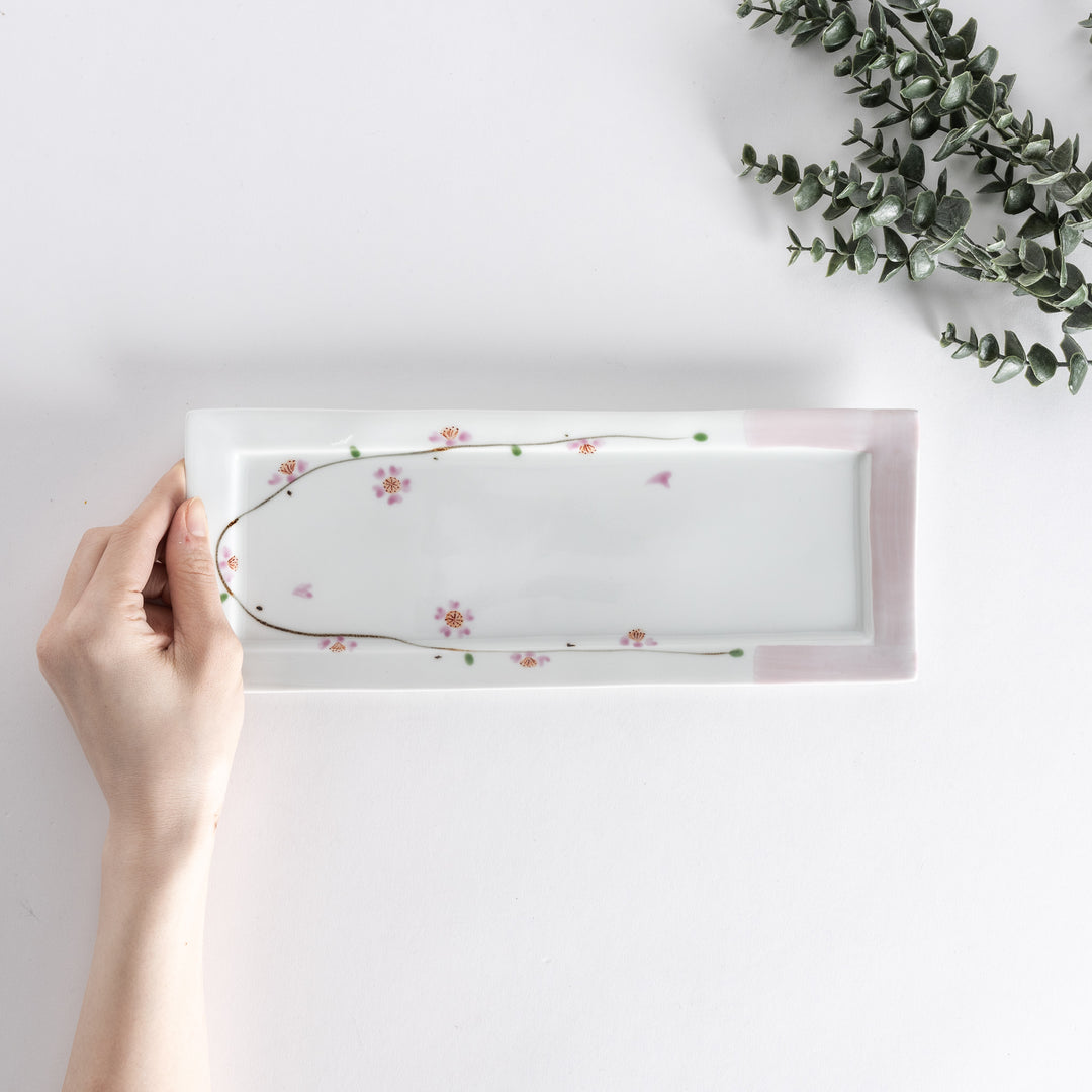 Hand holding a rectangular plate viewed from above, showcasing the delicate cherry blossom pattern with pink accents.