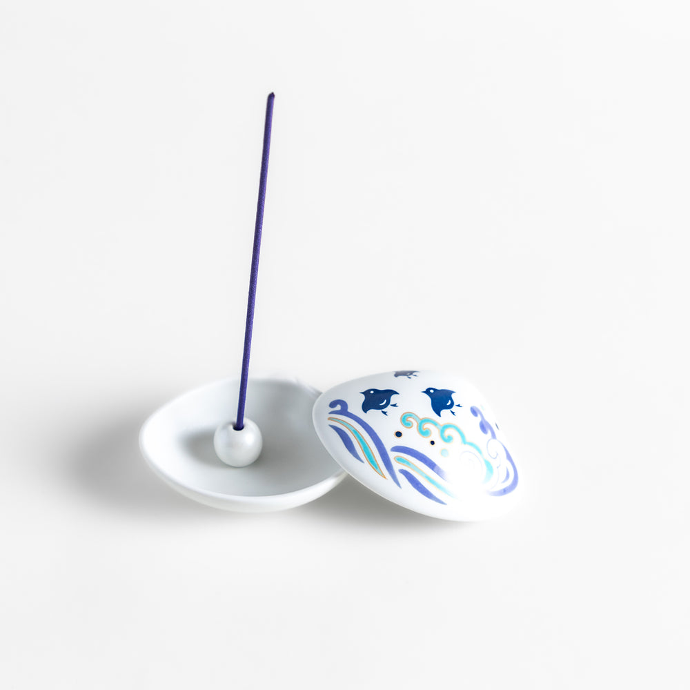 White ceramic incense holder shaped like a seashell, featuring an elegant blue wave and seabird motif, with a small rounded holder inside for secure incense placement.