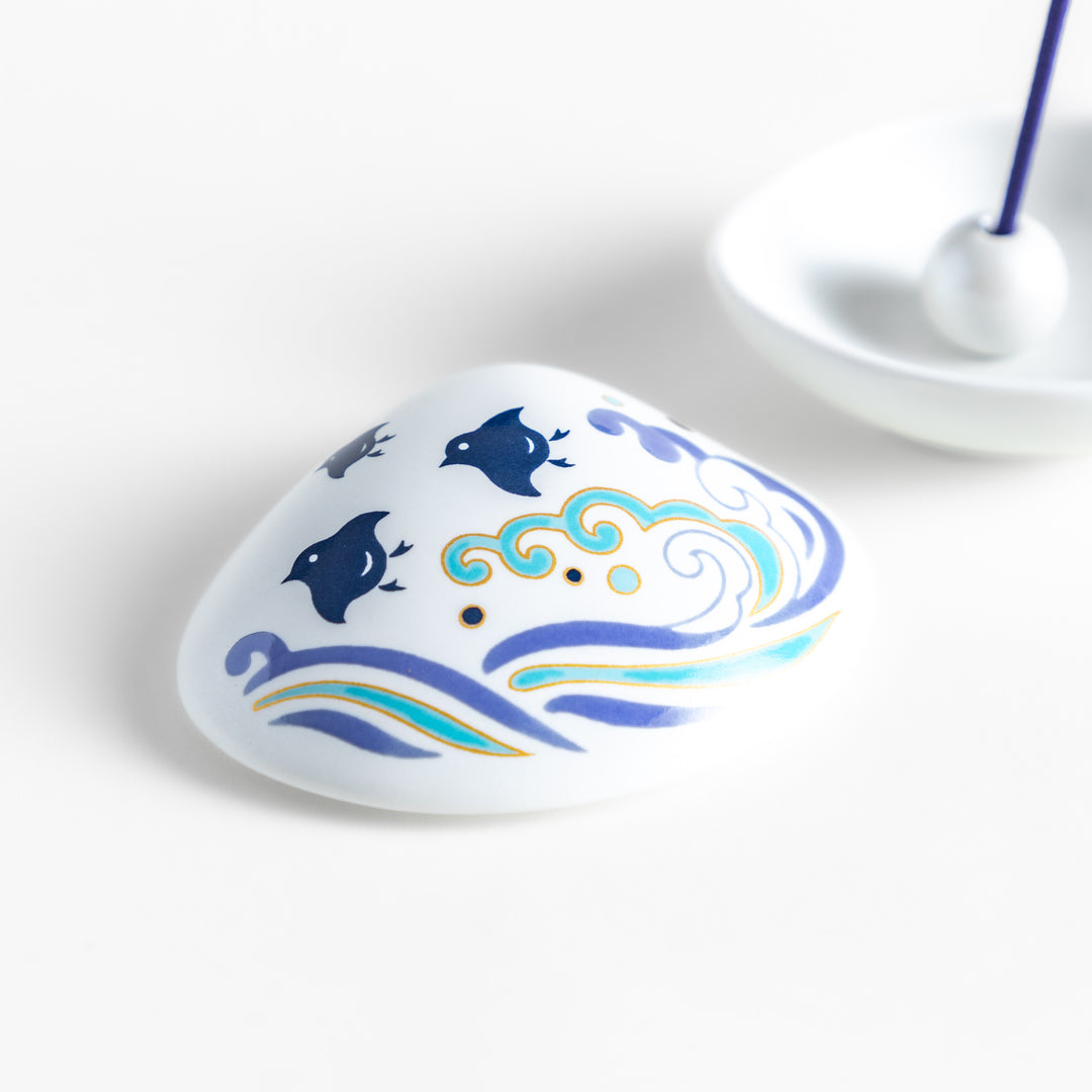 White ceramic incense holder shaped like a seashell, featuring an elegant blue wave and seabird motif, with a small rounded holder inside for secure incense placement.